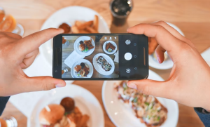 Photo shows two hands holding a cell phone poised to take a photo of a table of food.